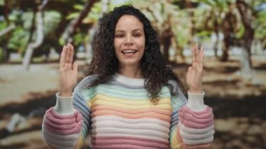 Parkta, açık havada, renkli bir süveter giymiş gözleri kapalı gülümseyen, ağaçlarla çevrili, güneşli bir ortamda el hareketi yapan bir kadın..