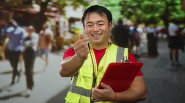 Güvenlik yeleği giyen genç adam, parmaklarıyla kalp hareketi yapıyor, kalabalık bir şehir arka planında gülümsüyor..