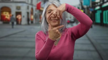 Gri saçlı son sınıf öğrencisi kadın, hareketli bir açık hava caddesinde, canlı ve sanatsal bir ifade sergilemek için ellerini kullanıyor.