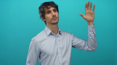 Açık mavi bir gömlek giymiş, mavi bir stüdyo duvarına karşı çıplak elini kaldıran ve portre çekimi bir avuç hareketi gösteren bir adam;.