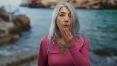 Gri saçlı bir kadın, deniz kenarındaki bir arka plana karşı ağzını şaşkınlıkla kapatıyor. Sahilde neşeli ve sıradan bir anı somutlaştırıyor..