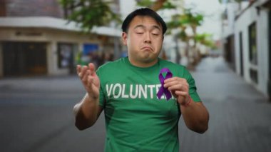 Gönüllü gömleği giyen genç bir adam, şehir sokaklarında mor bir kurdele tutuyor, hareketli bir şehirde farkındalık çabalarını simgeliyor..