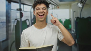 Sokak otobüsünün içinde pencerenin önünde otururken çeneye dokunurken açık kitap okuyan gözlüklü bir adam; tefekkür.