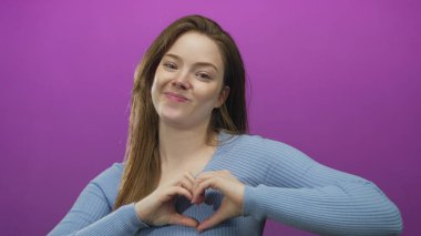 Mor stüdyoda elleri ile kalp şekli oluşturan genç beyaz kadın gülümsüyor; sevgi şefkat umut sevinç.