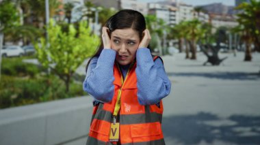 Turuncu yelekli kadın, arka planda yeşil ağaçlar olan kalabalık caddedeki gürültüden kulaklarını kapatıyor ve dışarıda rahatsızlığını dile getiriyor..