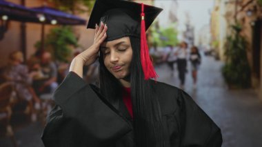 Mezuniyet şapkalı ve cüppeli genç İspanyol kadın arka planda bulanık insanlarla bir kafe terasında endişeli görünüyor..