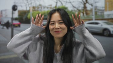 Uzun saçlı bir kadın sokakta yüz ifadeleri yapıyor. Bulanık arabaları ve yeşillikleri var. Açık hava ortamında çeşitli tepkiler gösteriyor..