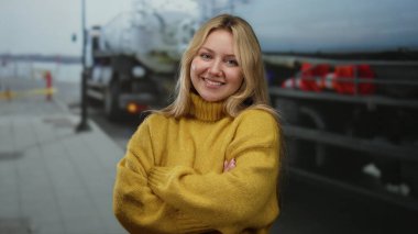 Şehirdeki inşaat sahasında kolları çaprazlanmış gülümseyen genç bir kadın donanım ve güvenlik engellerini gösteren bulanık bir geçmişe sahip sarı bir kazak giyiyor.