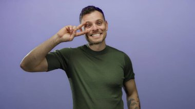 İki eliyle barış işareti yapan çekici genç bir adam canlı mor bir arka plana karşı neşeyle gülümsüyor. Dövmelerini ve günlük tarzını bir stüdyo ortamında sergiliyor.