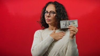 Gözlüklü orta yaşlı bir kadın Amerikan doları banknotunu kırmızı arka planda tutuyor, beyaz bir kazak giyiyor, odaklanmış bir ifadeyle kendini işaret ediyor..