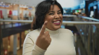 Canlı bir alışveriş merkezinde parmakları havada gülümseyen, kendine güveni ve neşe saçan, krem bir bluz giyen ve kapalı bir yerde, kalabalık dükkanların ortasında duran bir kadın..