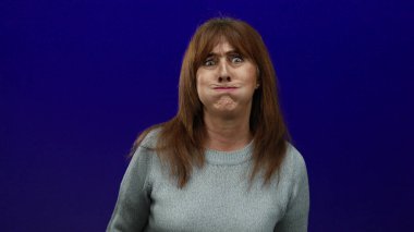 Katı mavi bir arka plana karşı komik suratlar yapan bir kadın, ellerinin yanaklarında olduğu mizah ve ifadeyi vurgulayan, tecrit edilmiş bir ortamda son sınıf İspanyol kadınını gösteren bir kadın..