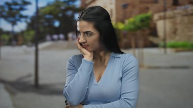 Genç İspanyol kadın güneşli bir sokakta dikiliyor. Mavi giyinmiş, ağaçların ve binaların arka planında huzurlu şehir hayatını yansıtıyor..