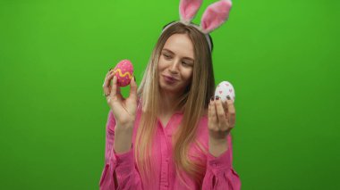 Tavşan kulaklı genç bir kadın, parlak yeşil bir arka planda paskalya yumurtalarını tutuyor. Renkli süslere neşeli bir şekilde gülümsüyor..
