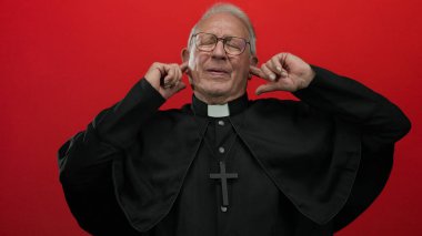 Siyah cüppeli kıdemli rahip ve kulakları kulaklarla kaplayan gözlüklü gözlüklü adam izole edilmiş kırmızı arka plana karşı hayal kırıklığı içinde..