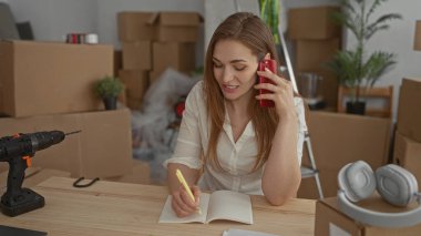 Kadın telefonda konuşurken inşaat masasında deftere yazı yazıyor; organizasyon geçiş beklentisi planlıyor.