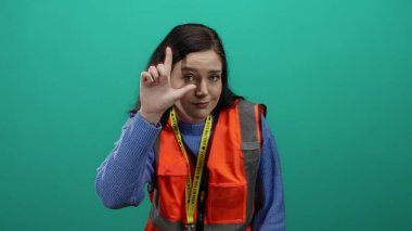 Gönüllü yelek giymiş esmer bir kadın yeşil bir zemine karşı ezik jestler yapıyor..