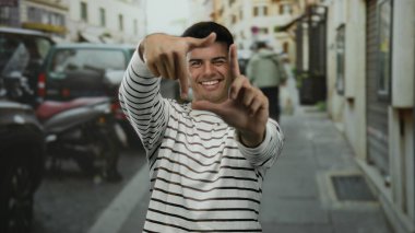 Çizgili tişörtlü genç adam gülümsüyor ve elleri ile şehir dışında canlı bir sokak arkaplanında çerçeveleniyor.