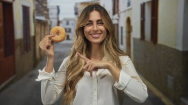 Genç sarışın kadın elinde tuttuğu donutu işaret ediyor güneş ışığında kaldırım taşı caddesinde; neşe aşeriyor.