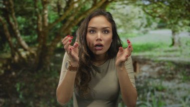 Yeşillik bir parkta sürpriz yapan, kırmızı tırnaklı İspanyol özelliklerini sergileyen doğa ve ağaçlarla çevrili, günlük kıyafetlerini giyen bir kadın..