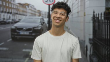 Gözlüklü ve park halindeki arabanın yanında gülümseyen bir adam ve kalabalık caddede beyaz bir bina; memnuniyet.