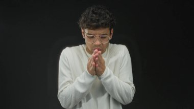 Siyah bir stüdyoda kollarını göğsünde kavuştururken gözlüklü ve beyaz kazaklı bir adam titriyor..