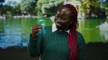 Parkta elinde mavi kurdeleyle gülümseyen bir kadın. Canlı yeşillik suya yansıyor. Açık havada barışçıl farkındalığı simgeliyor..