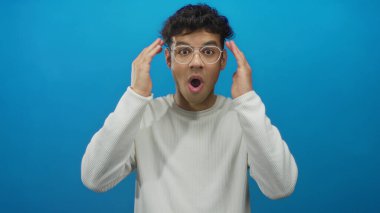 Beyaz eşofman altı giyen İspanyol genç adam ağzını açar ve mavi stüdyoda elleri yanaklarına bastırır..