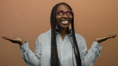 Gözlüklü bir kadın, kollarını açık bir şekilde yumuşak kahverengi bir arka plana yaslayarak neşeli bir şekilde gülümsüyor, örgülü saçlarını ve çizgili gömleğini parlaklaştırıyor..