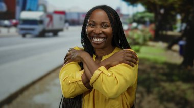 Bir kadın şehir caddesinde neşeli bir şekilde kendine sarılıyor, açık sarı elbisesi ve rahat tavırlarıyla şehir hayatının özünü yakalıyor..