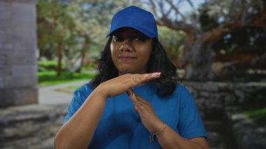 Mavi şapkalı ve t-shirtlü teslimatçı kadın parktaki ellerle jest yapıyor; dinginlik anı.