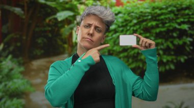 Kadın, orman ortamında yeşil yapraklara dayanan boş plastik kartı işaret ediyor; şüphecilik.