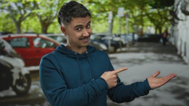 Mavi kapüşonlu genç İspanyol adam, arka planda park etmiş arabalar ve bulanık ağaçlarla dolu kalabalık bir şehir caddesinde avucunu göstererek baş parmağını kaldırıyor.