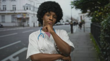 Steteskoplu kadın doktor, beyaz binalarla dolu bir caddede dururken çenesini uzatıyor ve kollarını kavuşturuyor..