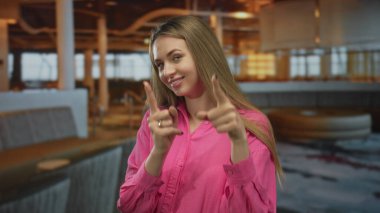 İçerideki bir restorana gülümseyen ve işaret eden bir kadın, kendine güvenen bir ifadeyle ve sıcak aydınlatma koşulları altında rahat bir atmosferle, parlak pembe bir gömlek ve uzun saçlı..