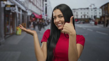 Kırmızı tişörtlü genç İspanyol kadın hareketli bir şehir caddesinde olumlu duygular ve açık havada kendine güvenen vücut dilini gösteriyor..