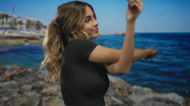 At kuyruklu kadın, parlak mavi gökyüzü altında kayalık deniz kıyısı tarafından esneme hareketinde çıplak kollarını uzatır; fitness gevşeme sükuneti.