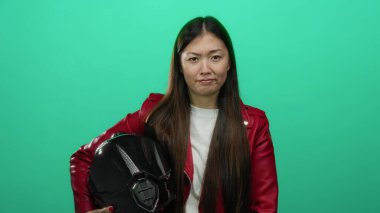 Kırmızı ceketli kadın yeşil arka planda miğfer tutuyor, güçlü bir ifade ve uzun saçlar sergiliyor, kendinden emin, şık bir hava yayıyor, modern, kentsel, kadın varlığını sembolize ediyor.