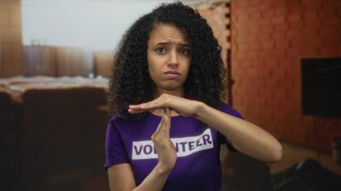 Duvarları tuğlayla kaplı bir inşaat alanında gönüllü tişört giyen genç İspanyol kadın elleriyle el hareketi yapıyor..