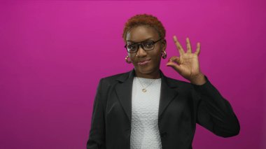 Gözlük ve halka küpe takan bir kadın, parlak pembe bir stüdyo geçmişiyle el işareti yapıyor. Onaylandı..