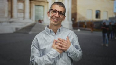 Gözlüklü ve elleri göğsünde gülümseyen bir adam arka planda tarihi bir binayla hareketli bir sokakta duruyor ve sıcak ve büyüleyici bir şehir manzarası yaratıyor..