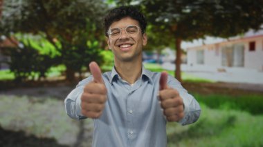 Genç İspanyol adam, yapraklı ağaçların altındaki güneşli yeşil parkta gülümserken iki kez baş parmak işareti yapıyor..