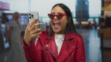 Kırmızı ceketli ve güneş gözlüklü bir kadın havaalanı terminalinde selfie çekiyor..