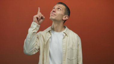 Kırmızı duvara yaslanmış bej gömlekli bir adam, düşünceli bir ifadeyle yukarıya doğru bakıyor. İzole edilmiş bir alanda fikri ya da ilhamı simgeliyor..