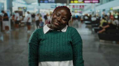 Havaalanı terminalinde oturan yeşil kazaklı kadın, mutlu bir ifadeyle, kalabalık gezginlerle çevrili, kapalı bir havaalanı ortamında..