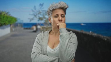 Gri kapüşonlu kısa gri saçlı kadın, sokakta çene jesti el ele durur; sessiz tefekkür.