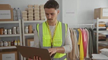 Yansıtıcı yelek giyen genç bir adam bir hayır kurumunda dizüstü bilgisayar üzerinde çalışıyor. Etrafı yiyecek ve giysi dolu raflarla çevrili. Gönüllü faaliyetlerde bulunuyor..