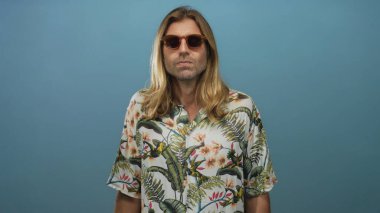 Uzun sarı saçlı, güneş gözlüğü takan ve tropikal gömlekli bir adam stüdyo odasında güven içinde poz veriyor..