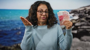 Elinde İzlanda kronu olan orta yaşlı bir kadın deniz manzarası üzerinde duruyor, sakin bir plaj zeminine karşı ekonomik temaları vurguluyor..