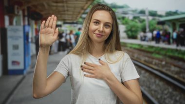 Genç bir kadın, tren istasyonunda, açık havada, arka planda demiryolu raylarıyla el ele tutuşuyor, gülümsüyor ve beyaz gömlek giyiyor, bulanık kalabalıklar ve yeşillikler içinde..